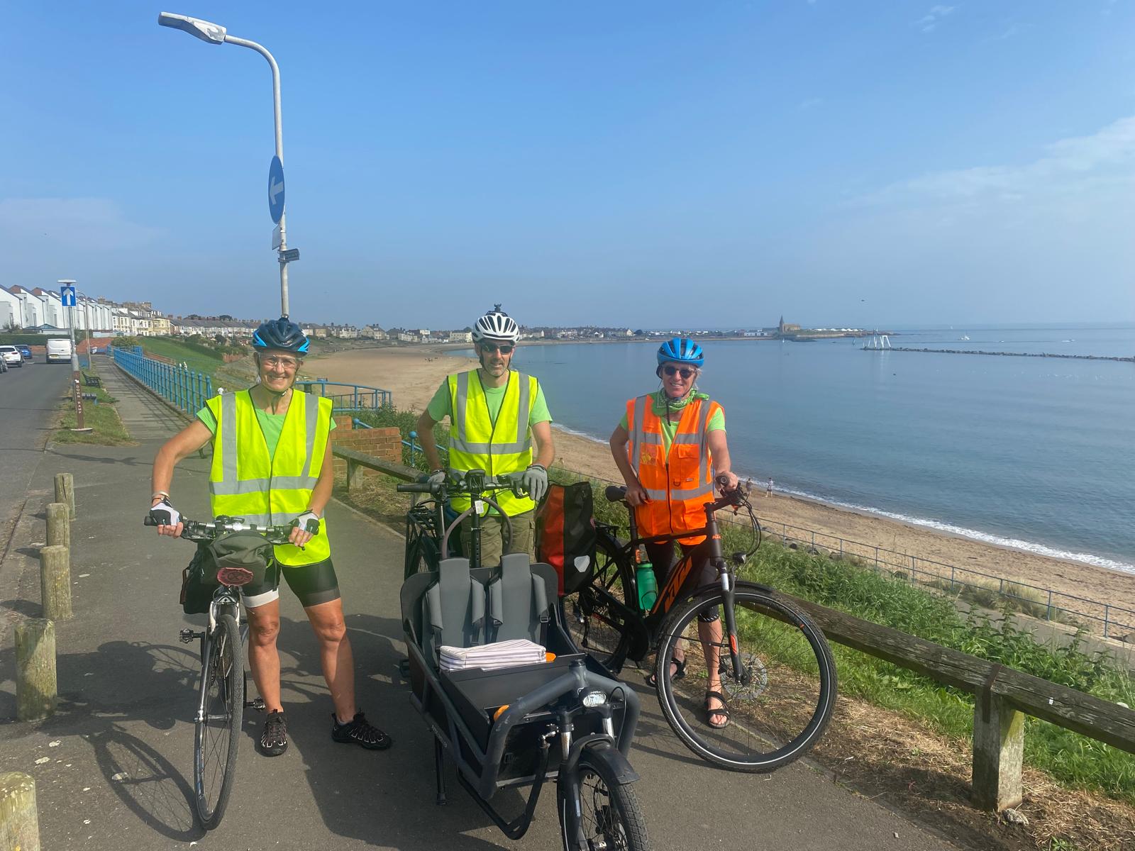 Three cyclists in high viz standing with their bikes on a road by the coast